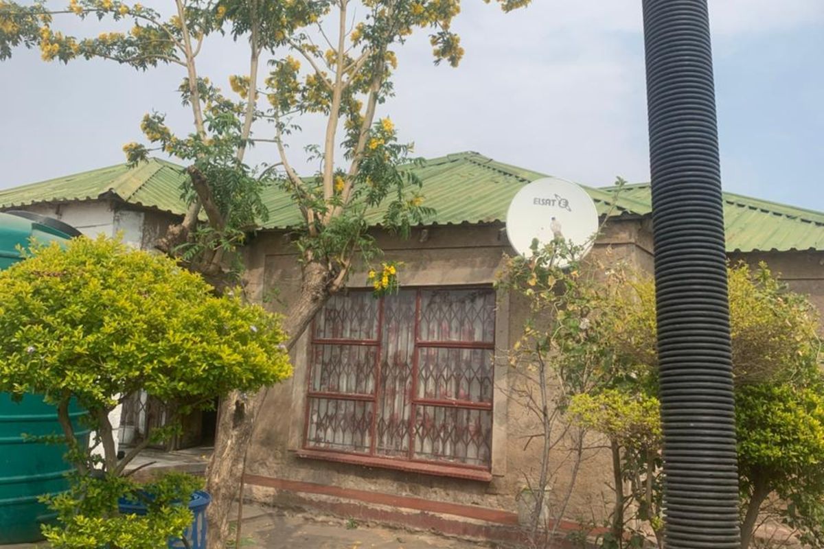 Exterior view of the property featuring a green metal roof, security-barred windows, and a satellite dish, set within a garden area.