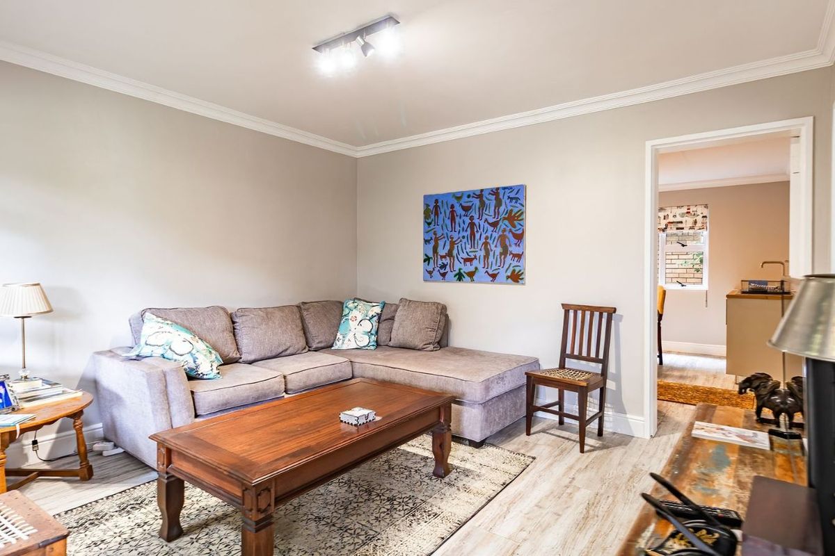 A comfortable living room featuring neutral wall tones, crown molding, and light-toned wood-look flooring, with an open doorway leading to an adjacent kitchen area.