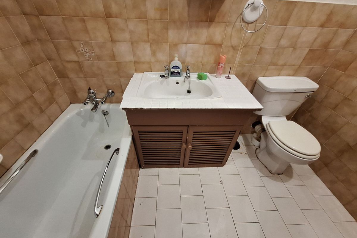 A dated bathroom featuring a standard bathtub, a vanity sink unit, and a toilet, finished with brown wall tiles and white floor tiles.