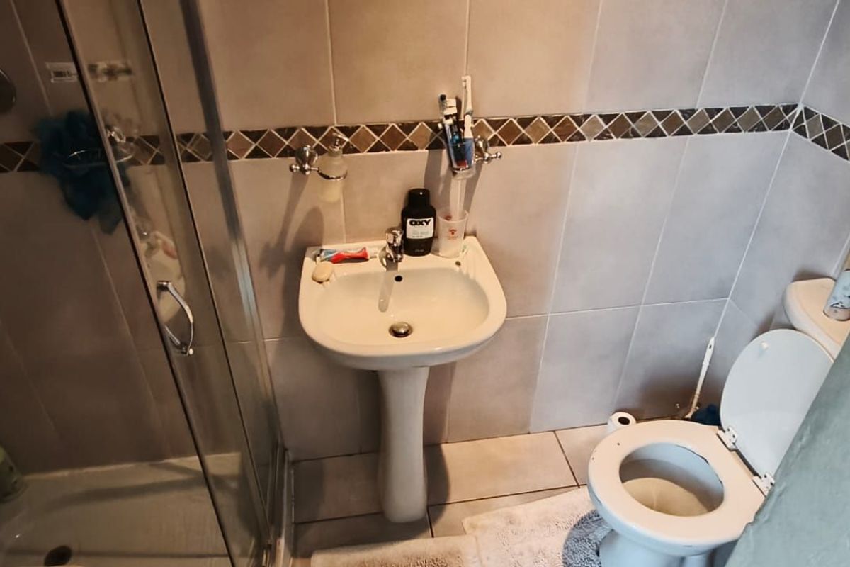 A compact bathroom featuring a pedestal sink, a glass-enclosed shower, and a standard toilet with neutral wall and floor tiling.