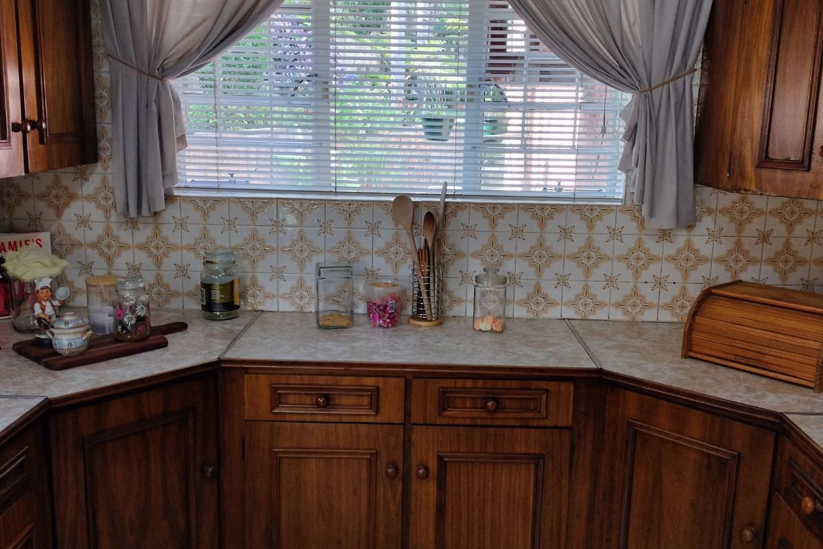 A dated kitchen featuring traditional wooden cabinetry, patterned wall tiles, and a central window with blinds, offering a classic aesthetic ready for modernization.