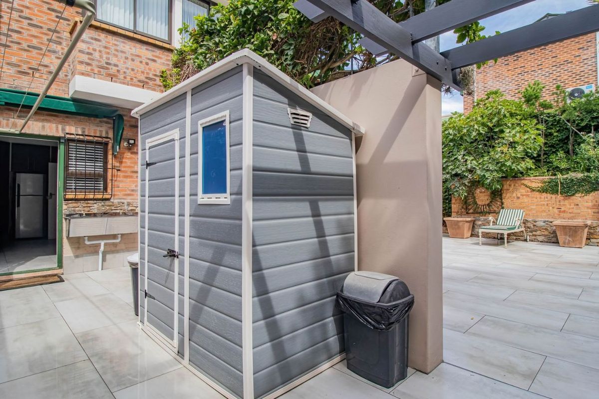 A paved rear courtyard featuring a grey plastic storage shed, a built-in outdoor wash basin, and a dark timber pergola structure.
