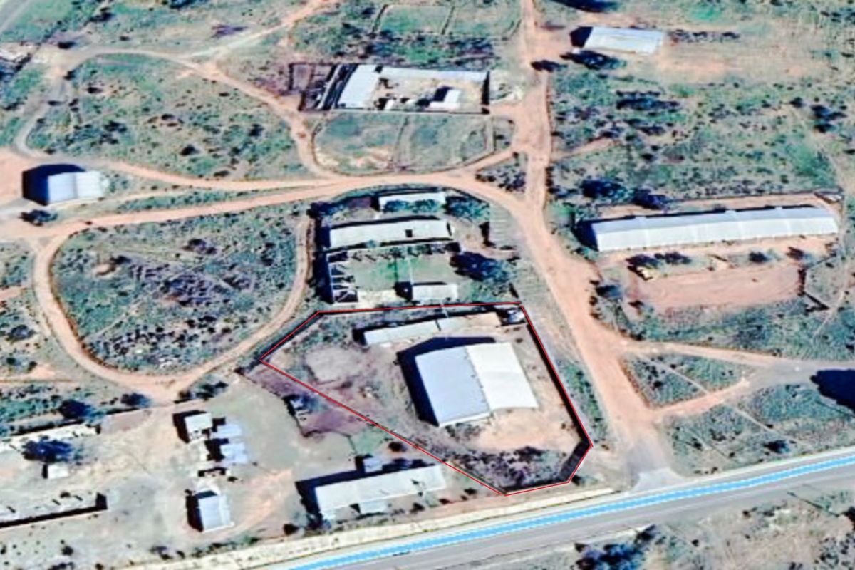 Aerial view of an industrial property site featuring multiple warehouse structures, gravel access paths, and perimeter fencing in a rural setting.