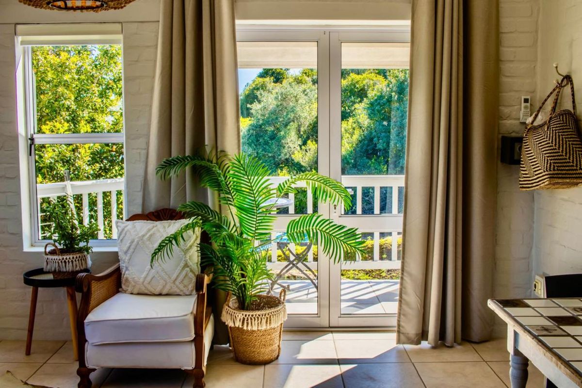 A bright living area featuring tiled flooring, white painted brick walls, and double glass doors opening onto a private balcony with garden views.