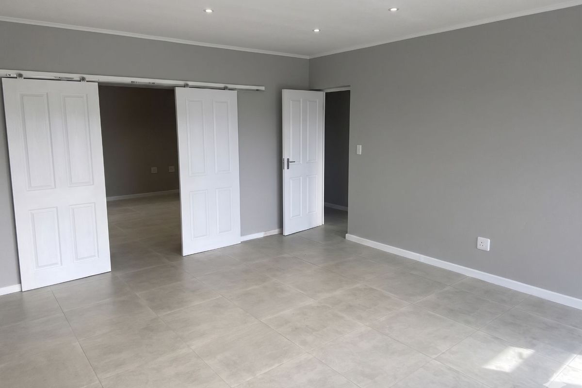 A spacious, neutral-toned living area featuring large format floor tiles, recessed downlighting, and white panelled sliding and standard doors.