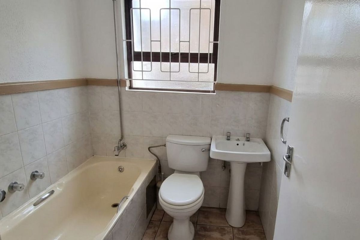 A compact bathroom featuring a bathtub, pedestal basin, and toilet, with tiled walls and a security-grille window.