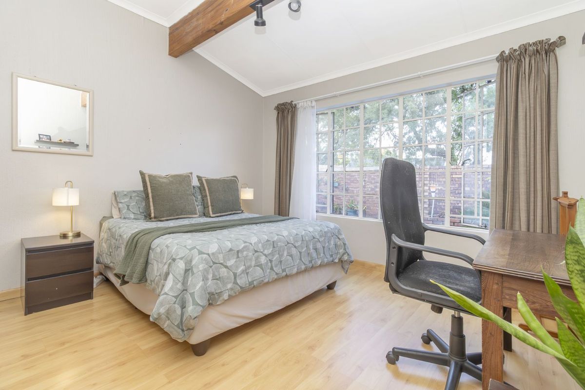 A bedroom featuring light wood-look flooring, a vaulted ceiling with an exposed timber beam, and large windows providing natural light and garden views.
