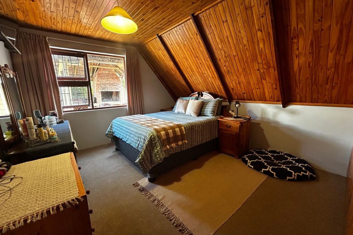 A cozy bedroom featuring a sloped timber-paneled ceiling, carpeted flooring, and a window overlooking an outdoor patio area.