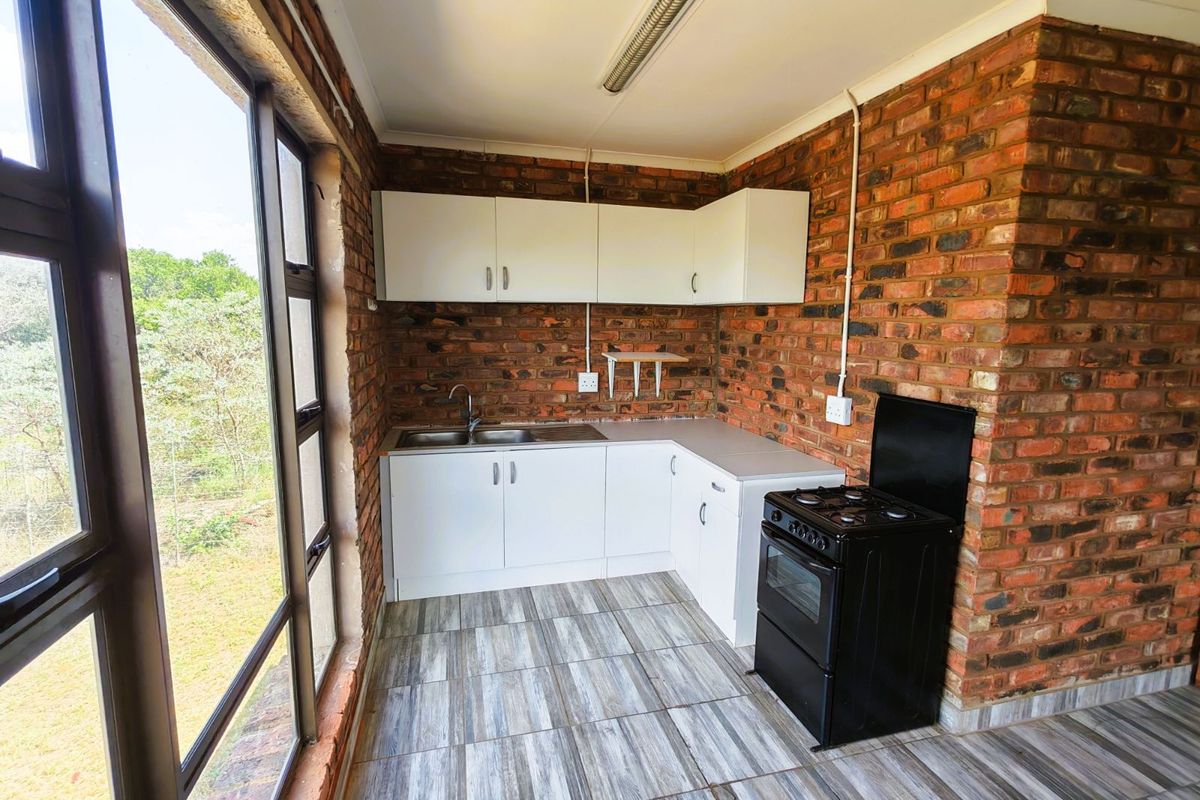 A compact kitchen featuring exposed brick walls, white cabinetry, a stainless steel sink, and a freestanding stove, with large windows overlooking the farm landscape.