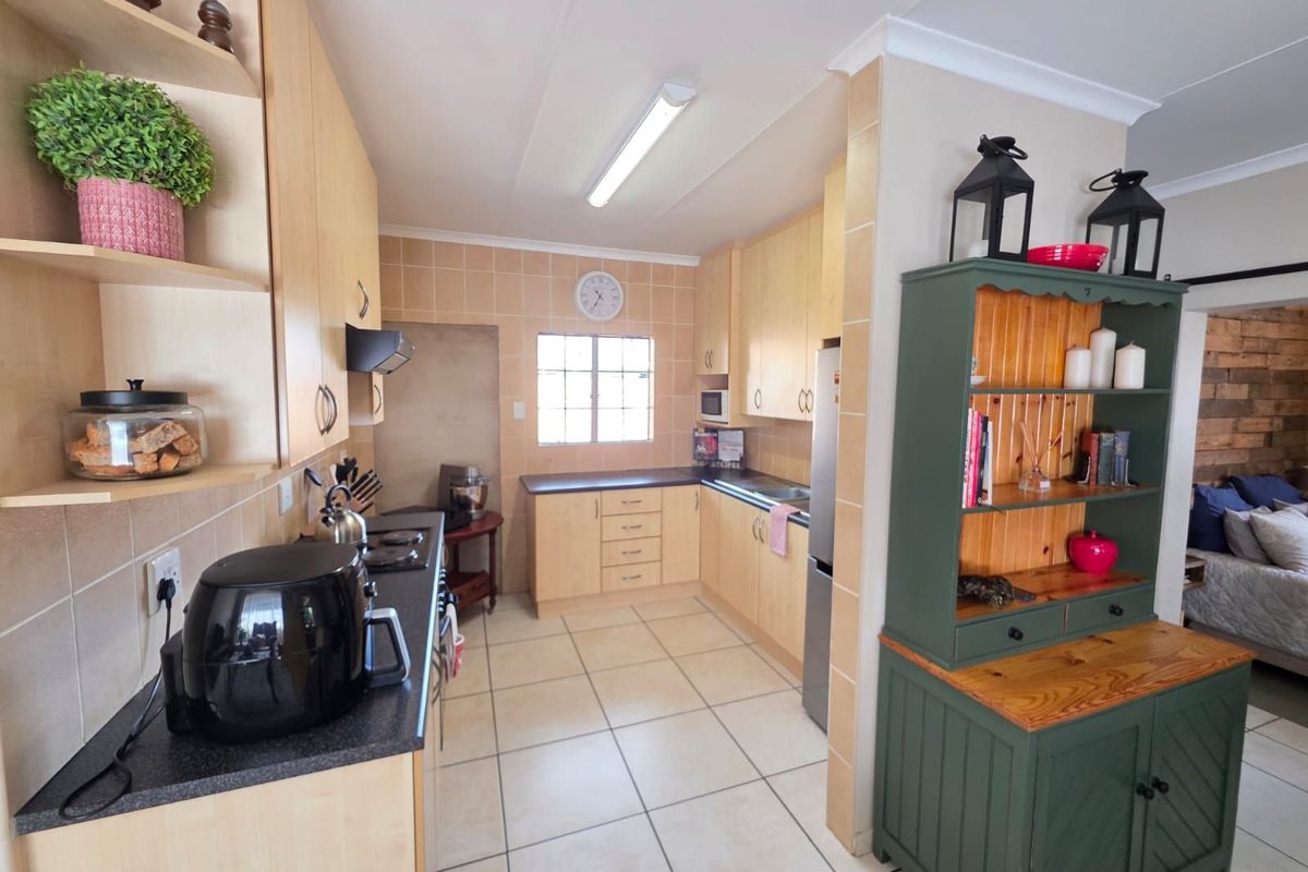 A functional kitchen featuring light wood cabinetry, dark countertops, tiled splashbacks, and tiled flooring, with an open-plan layout connecting to the living area.