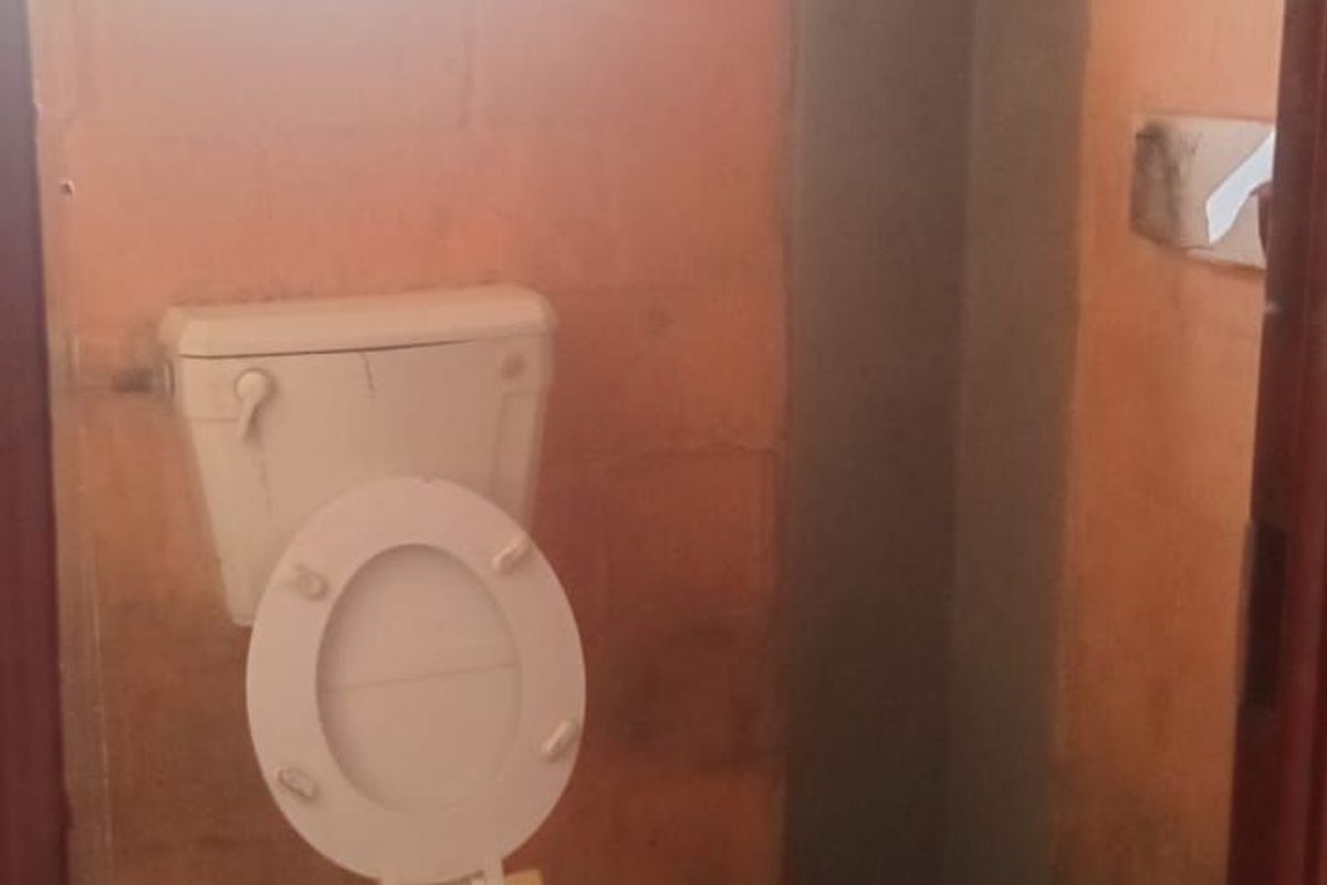 A small, basic bathroom featuring a standard toilet, unfinished block walls, and tiled flooring, requiring significant cosmetic updates and maintenance.