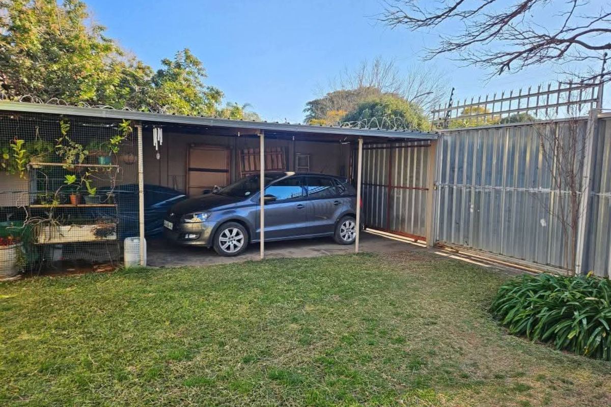 A functional outdoor area featuring a covered carport structure, metal perimeter fencing, and a grassed garden space.