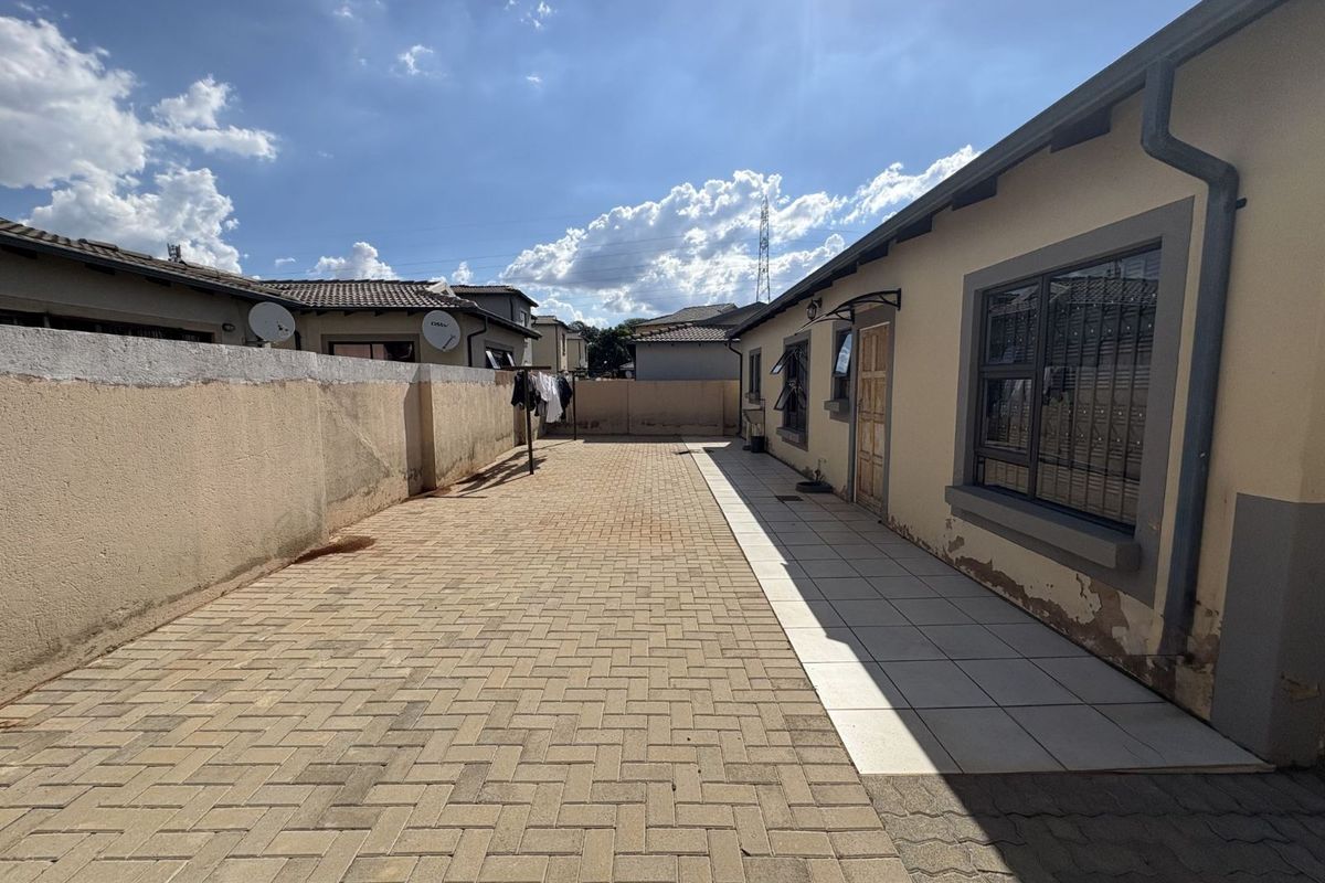 A paved rear exterior area featuring a tiled walkway along the side of the house and a perimeter boundary wall.