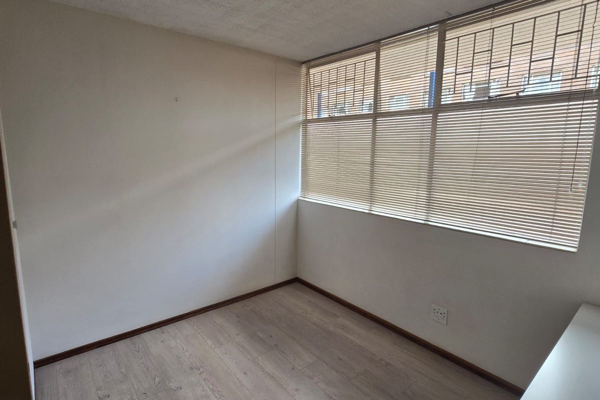 A bright, empty bedroom featuring light wood-look laminate flooring, large windows with fitted blinds, and neutral wall finishes.