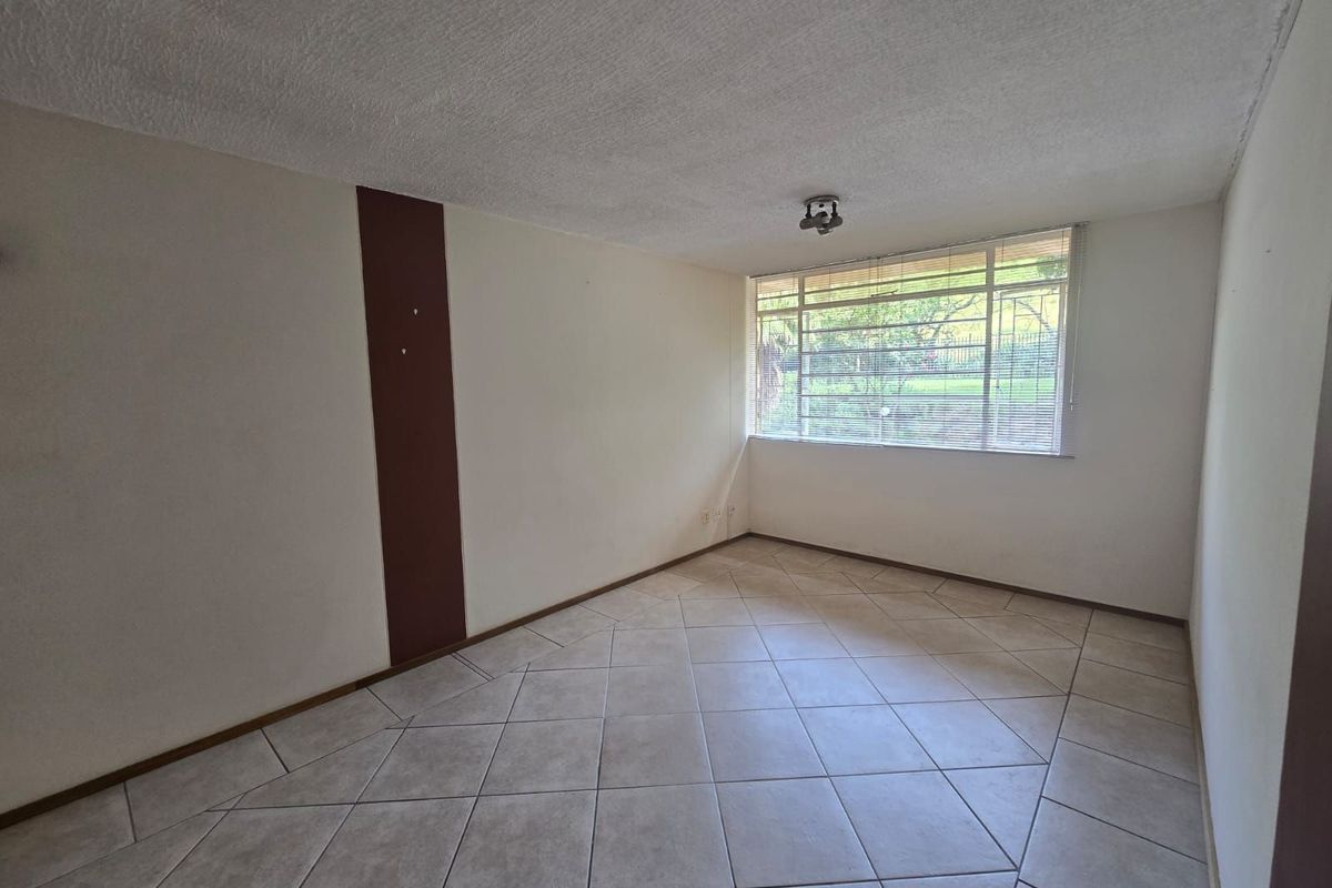A bright, empty living room featuring neutral ceramic floor tiles, a large window with fitted blinds, and a decorative wall accent.
