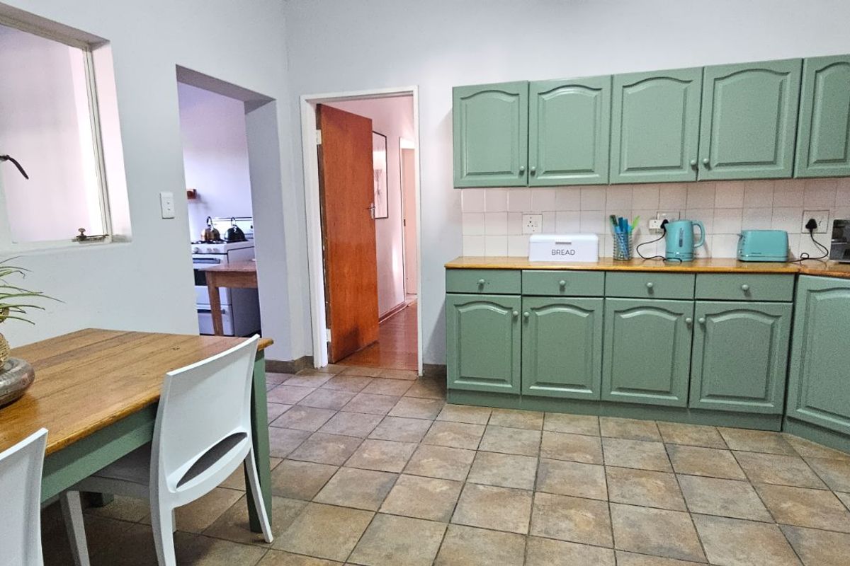 A functional kitchen featuring sage green cabinetry, tiled flooring, and a tiled backsplash, with an adjacent dining area and access to a scullery.