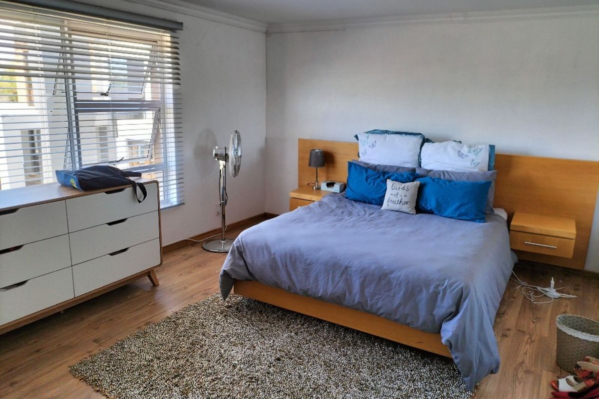 A well-lit bedroom featuring wood-look laminate flooring, a built-in wooden headboard with integrated bedside tables, and large windows with venetian blinds.