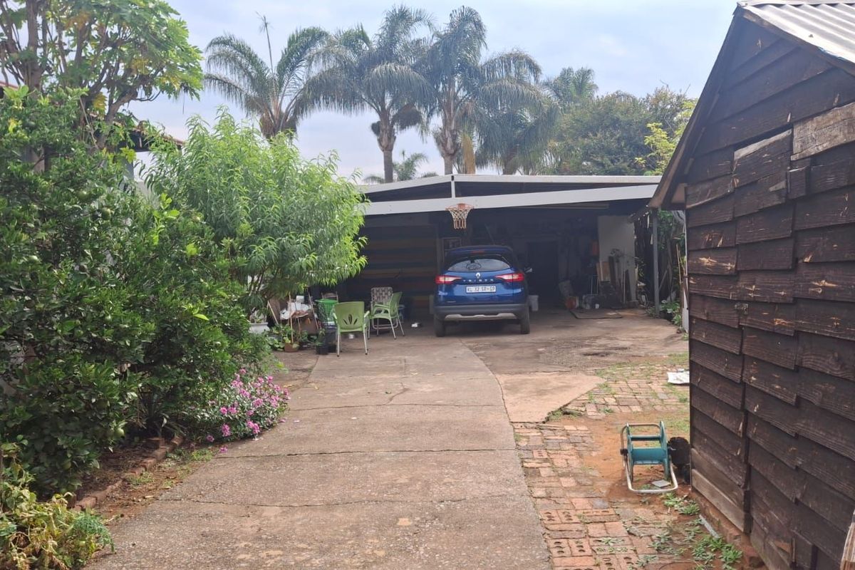 A view of the rear property area featuring a concrete driveway, a covered carport structure, and a timber-clad storage shed in the garden.