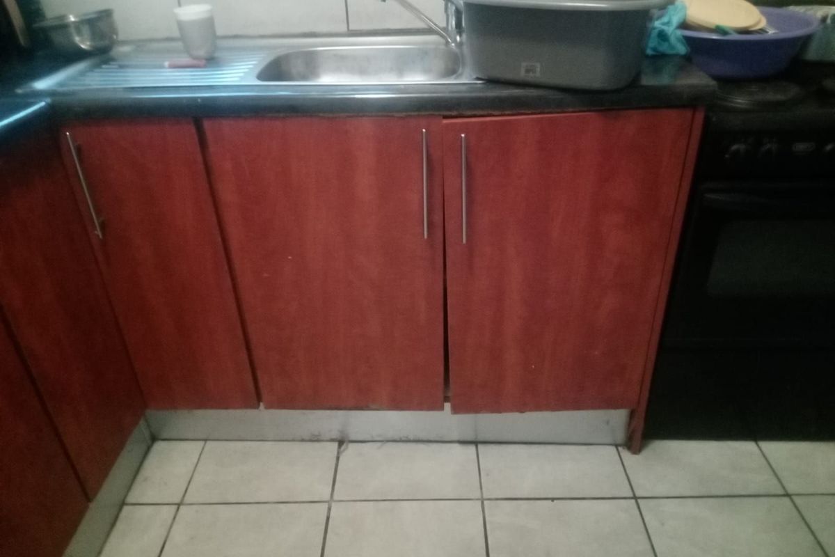 A compact kitchen area featuring red-toned cabinetry, a stainless steel sink, and tiled flooring, showing signs of wear and tear.