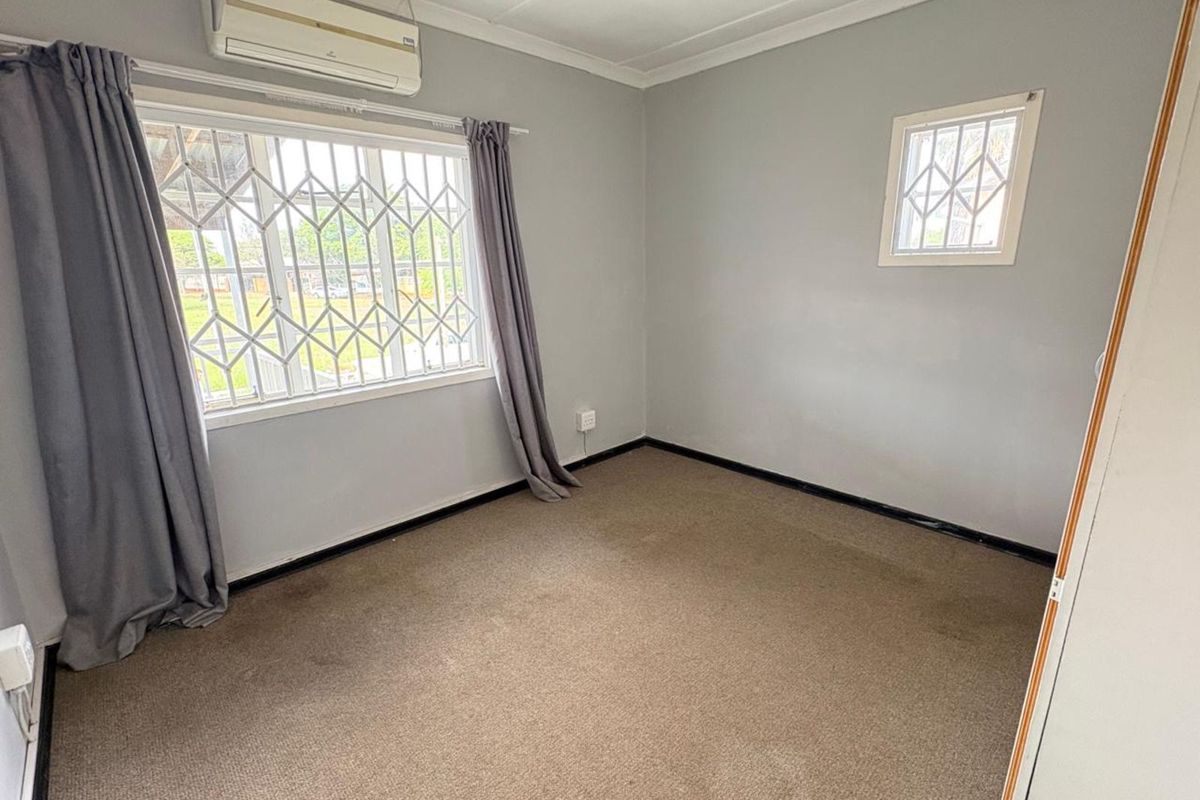 A neutral-toned bedroom featuring carpeted flooring, a wall-mounted air conditioning unit, and security bars on the windows.