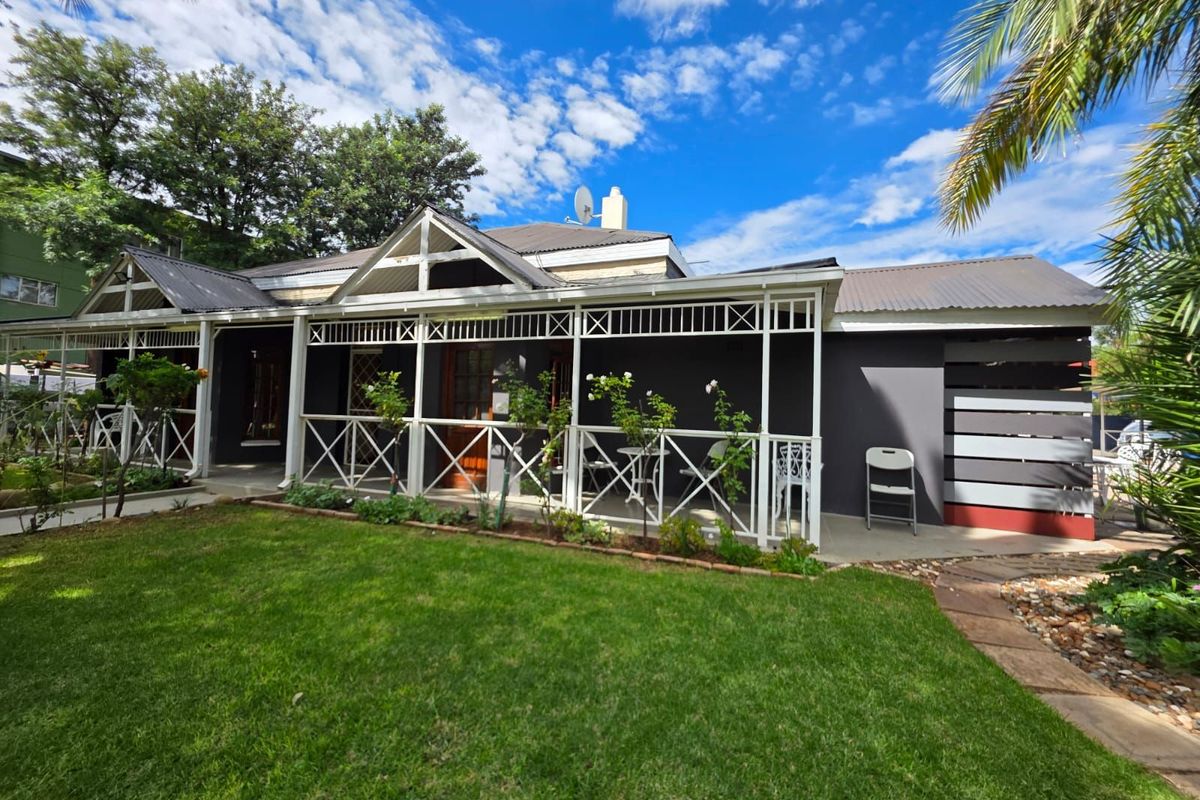 A well-maintained guest house exterior featuring a covered veranda with white lattice detailing, a manicured lawn, and a paved garden path.