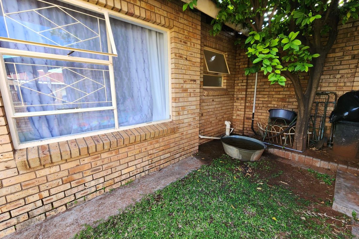 A view of the exterior rear brick wall featuring a window with security bars, a small garden area, and exposed utility plumbing.
