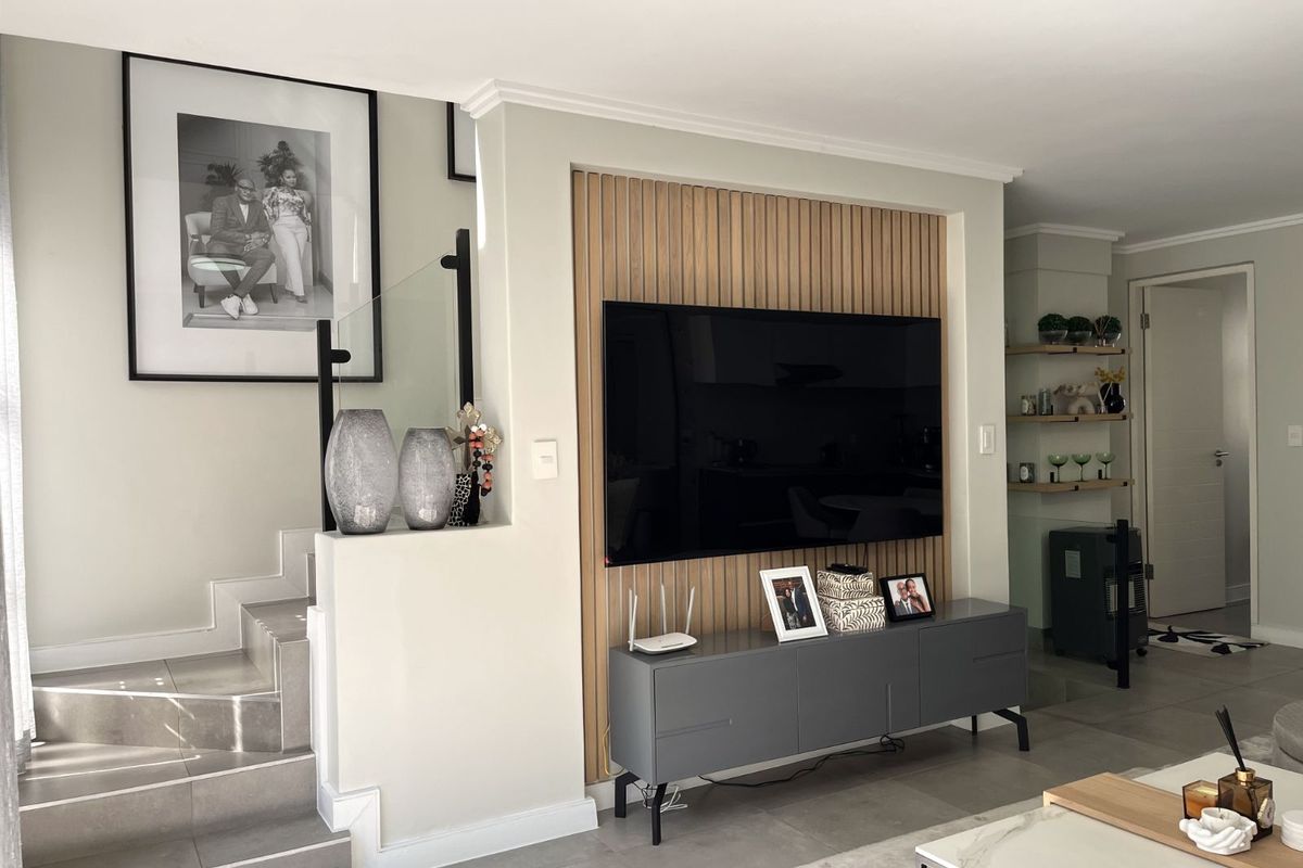 A modern living room featuring a decorative timber slat feature wall, tiled flooring, and an integrated staircase with a glass balustrade.