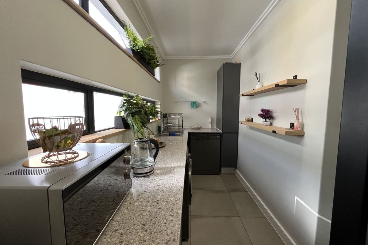 A modern, narrow galley-style kitchen featuring stone countertops, dark cabinetry, integrated appliances, and floating timber shelving under clerestory windows.