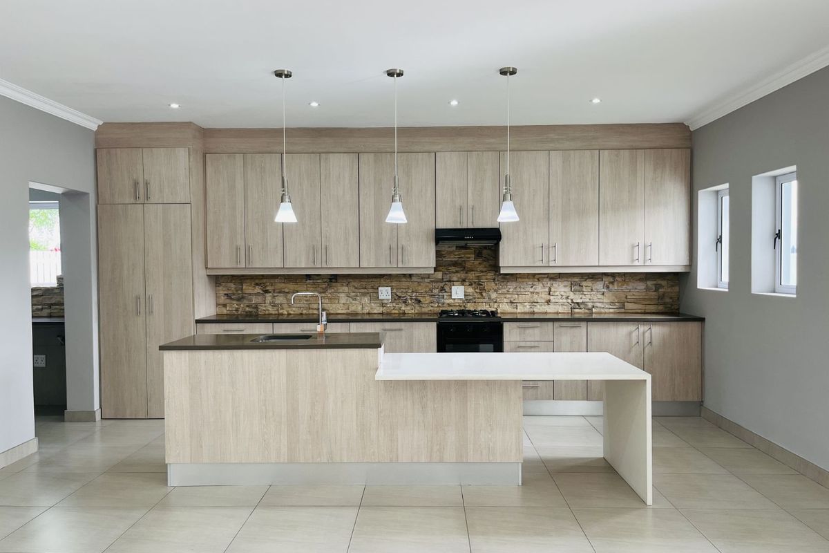 Modern kitchen featuring light wood-grain cabinetry, stone-cladding backsplash, dark countertops, a central island with integrated sink, and sleek pendant lighting.
