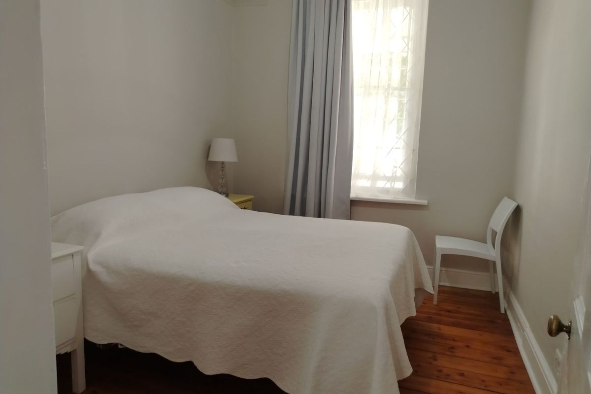 A simple bedroom featuring original timber flooring, neutral wall finishes, and a window with curtain treatments.