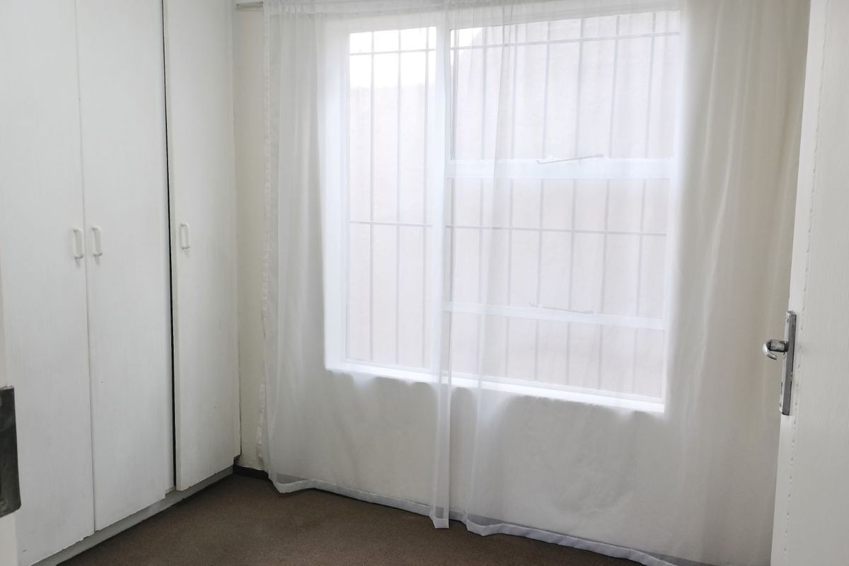 A simple bedroom featuring built-in cupboards and a window with security bars, offering a functional and neutral space.