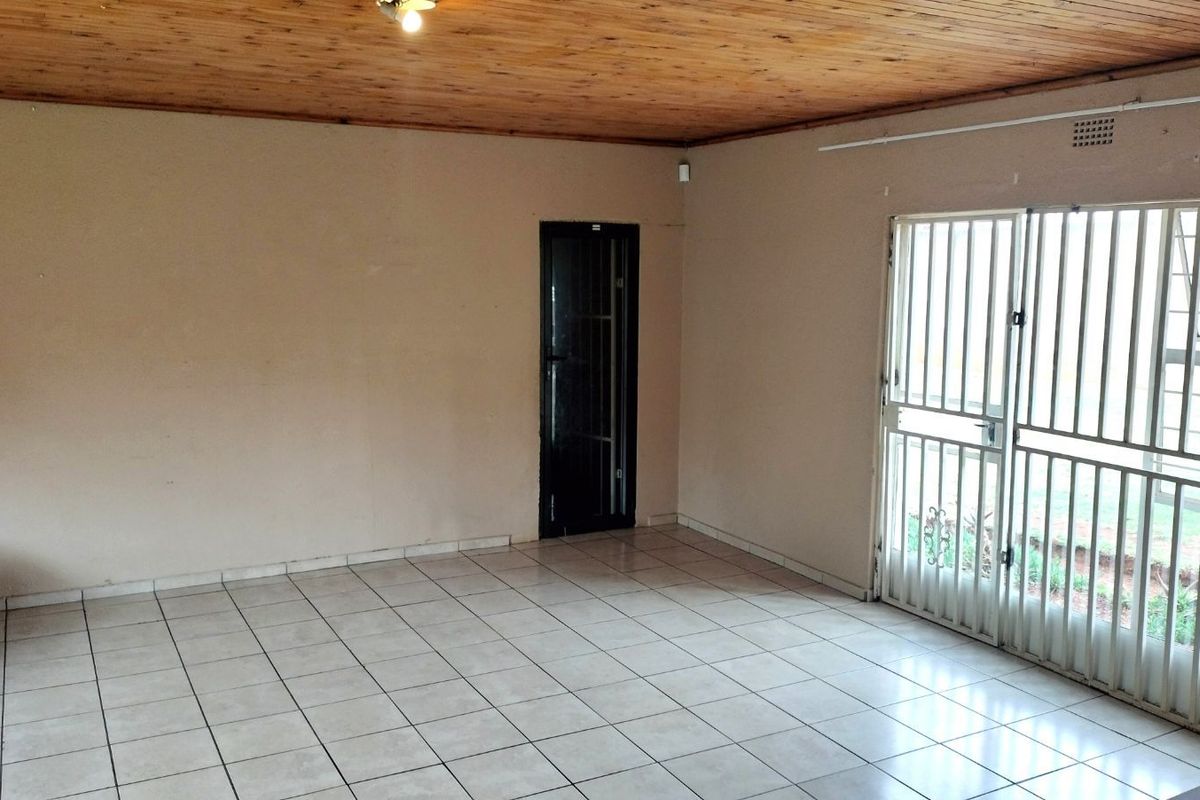 An empty living room featuring tiled flooring, a wooden panelled ceiling, and a security gate leading to the exterior.