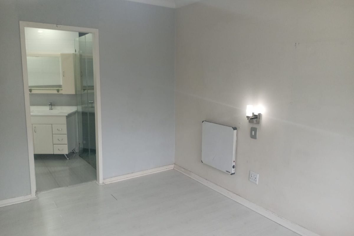 A bright bedroom featuring light-toned flooring, a wall-mounted heater, and an adjoining en-suite bathroom with a vanity and glass shower enclosure.