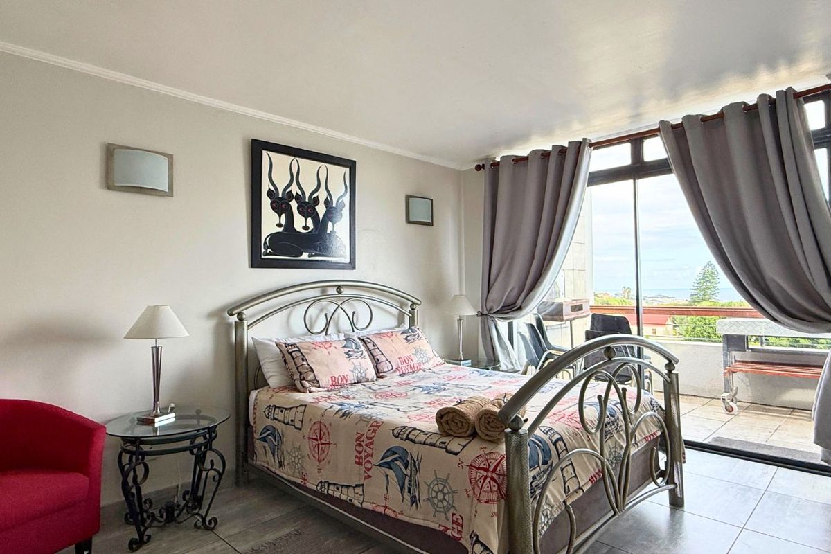 A well-lit bedroom featuring tiled flooring, wall-mounted lighting, and large sliding glass doors that open onto a private balcony with coastal views.