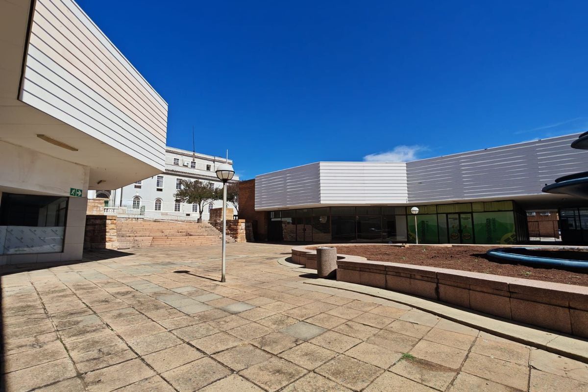 An outdoor commercial courtyard area featuring paved surfaces, raised garden beds, and modern building facades with large glass storefronts.