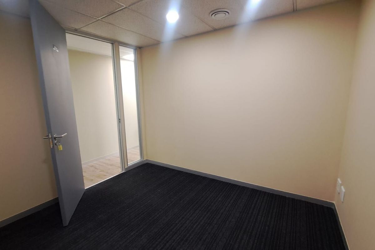 A clean, empty commercial office space featuring dark carpeting, a suspended ceiling with recessed lighting, and a glass-paneled door entry.