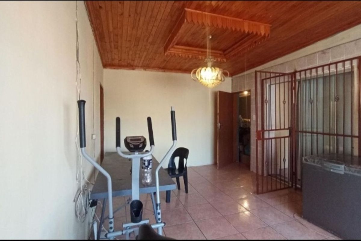 A multi-purpose living area featuring tiled flooring, a wooden panelled ceiling with a central light fixture, and a security gate leading to an exterior space.