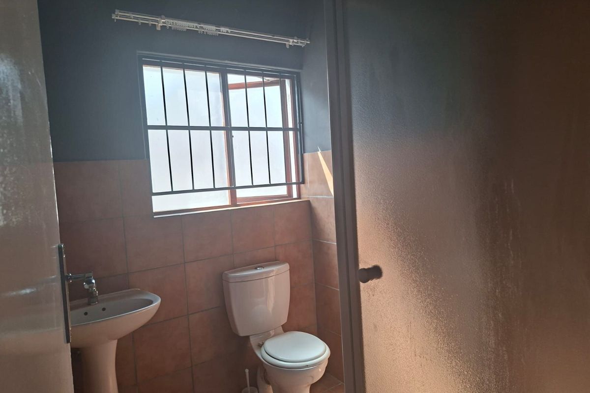 A functional bathroom featuring a pedestal basin, standard toilet, and a window with security bars, finished with terracotta-style wall tiles.
