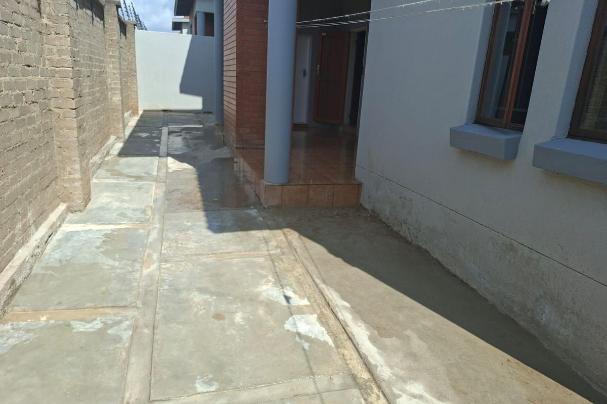 A side exterior view of the property featuring a concrete walkway, brick pillar detail, and perimeter boundary wall with security fencing.