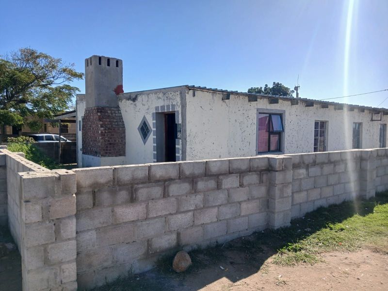 Front exterior view of a single-story house featuring a prominent brick chimney and a concrete block boundary wall, showing signs of significant exterior wear.