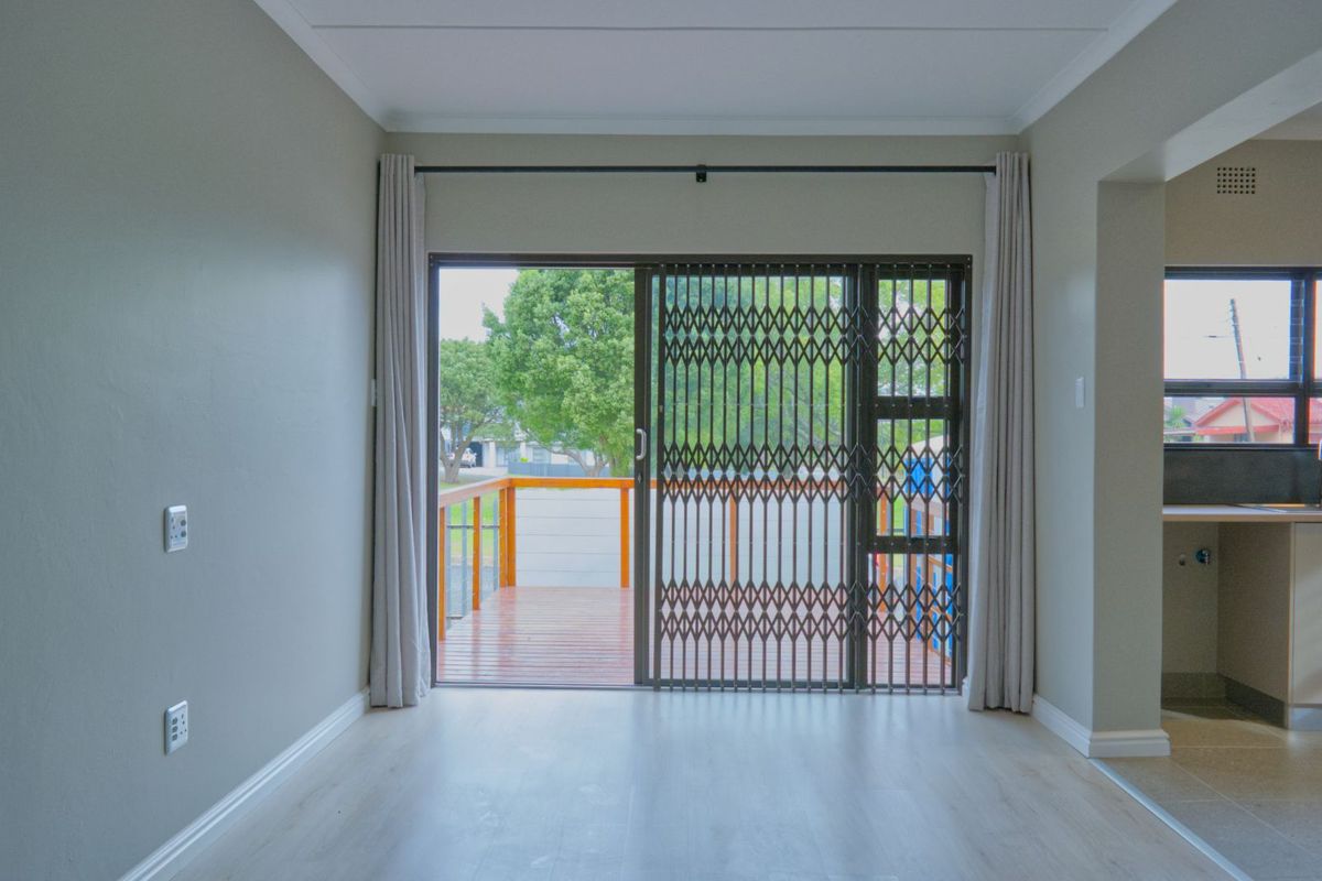 Bright living area featuring light-toned flooring, neutral walls, and a sliding glass door with security gate opening onto a wooden deck.