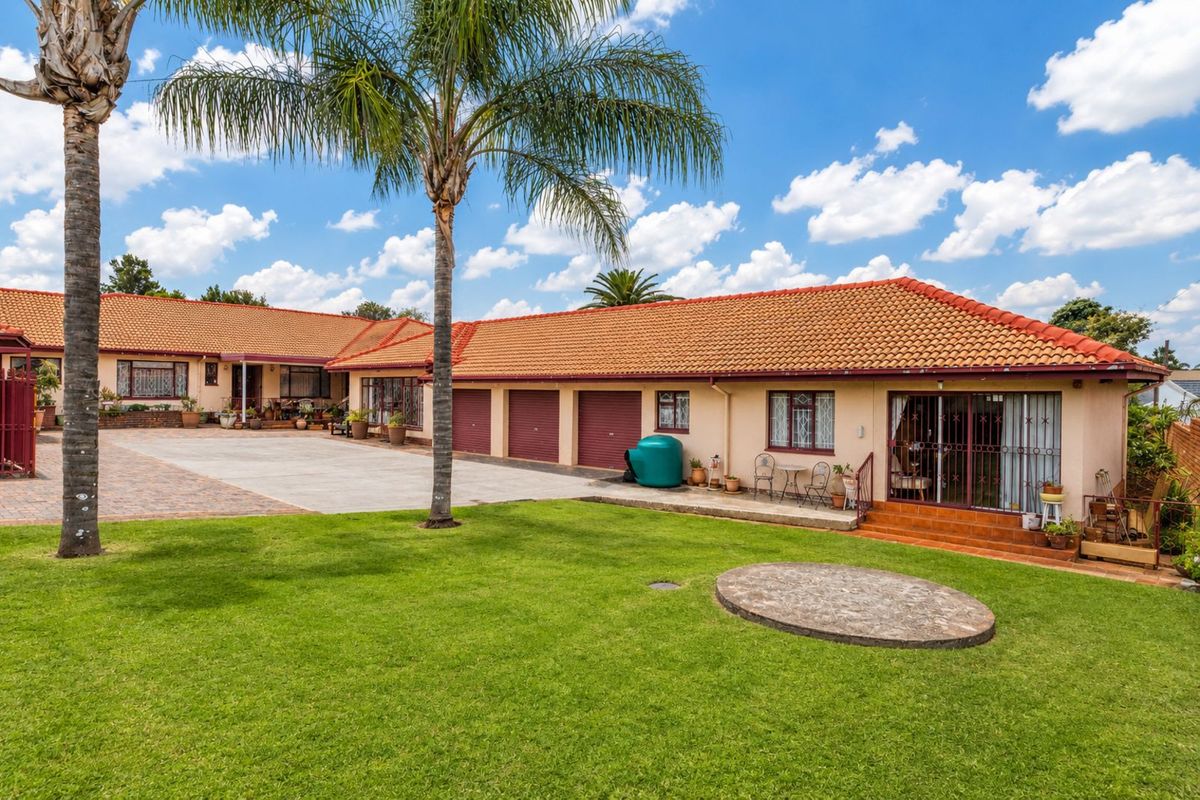 Spacious rear exterior view featuring a large lawn, paved courtyard, multiple garage doors, and a tiled roof structure with patio access.