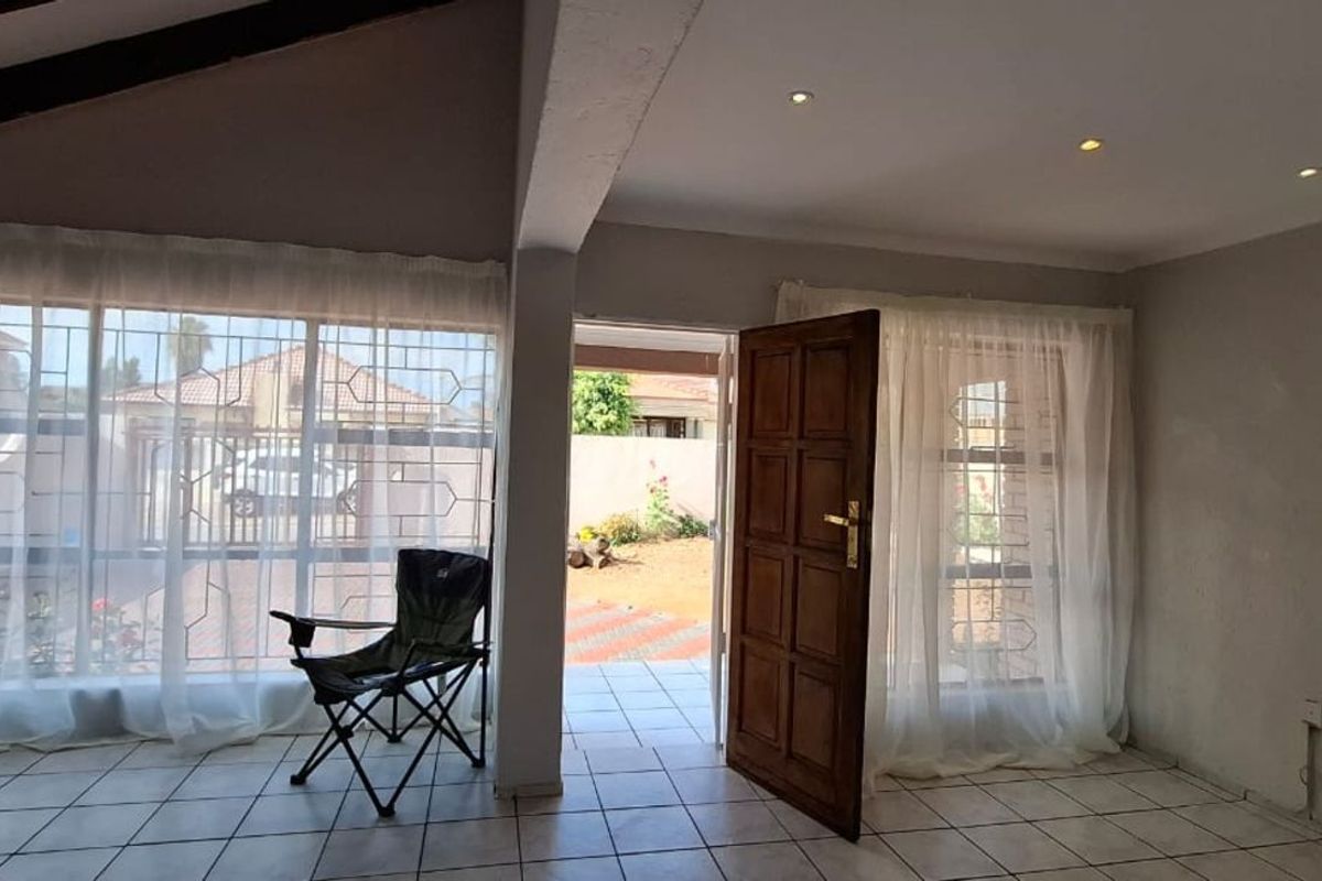 A bright living area featuring tiled flooring, exposed ceiling beams, and large windows with security bars, providing direct access to the outdoor patio.