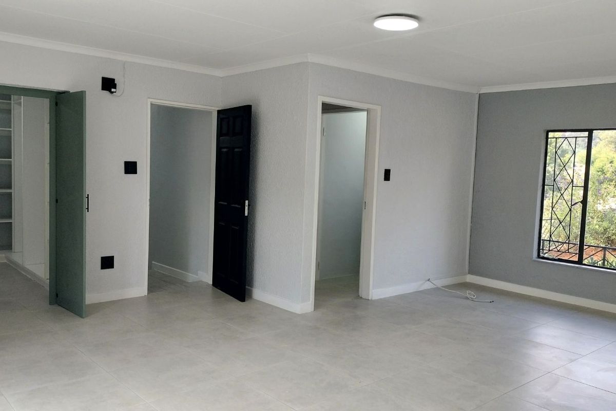 A bright, empty bedroom featuring neutral tiled flooring, built-in shelving, and a window with security bars overlooking the garden.