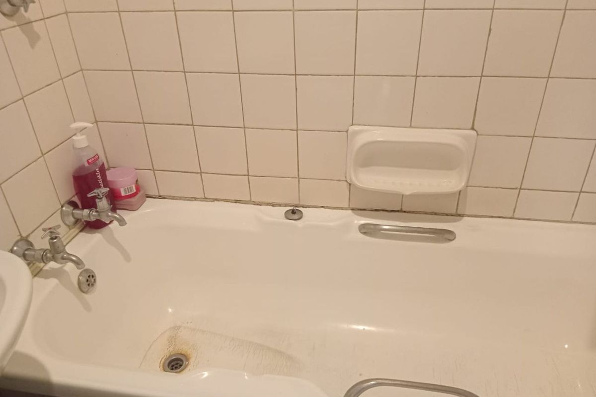 A dated bathroom featuring a standard white bathtub with wall-mounted taps, a built-in soap dish, and tiled splashback.