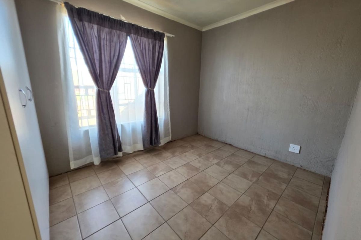 A simple, tiled bedroom featuring neutral walls, a built-in cupboard, and a window with security bars.