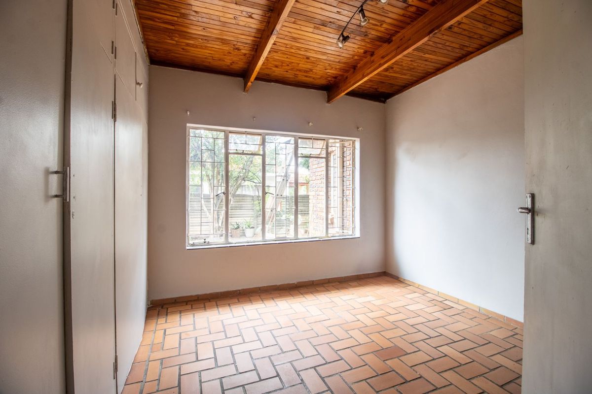 A bright bedroom featuring exposed timber ceiling beams, terracotta floor tiles, and built-in cupboards with a large window overlooking the garden.