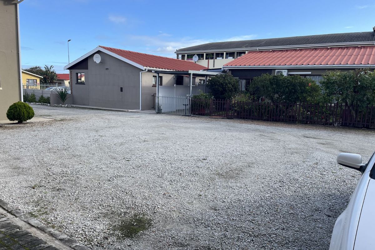 A view of the exterior rear area featuring a gravel parking space, metal fencing, and residential building structures with tiled roofs.