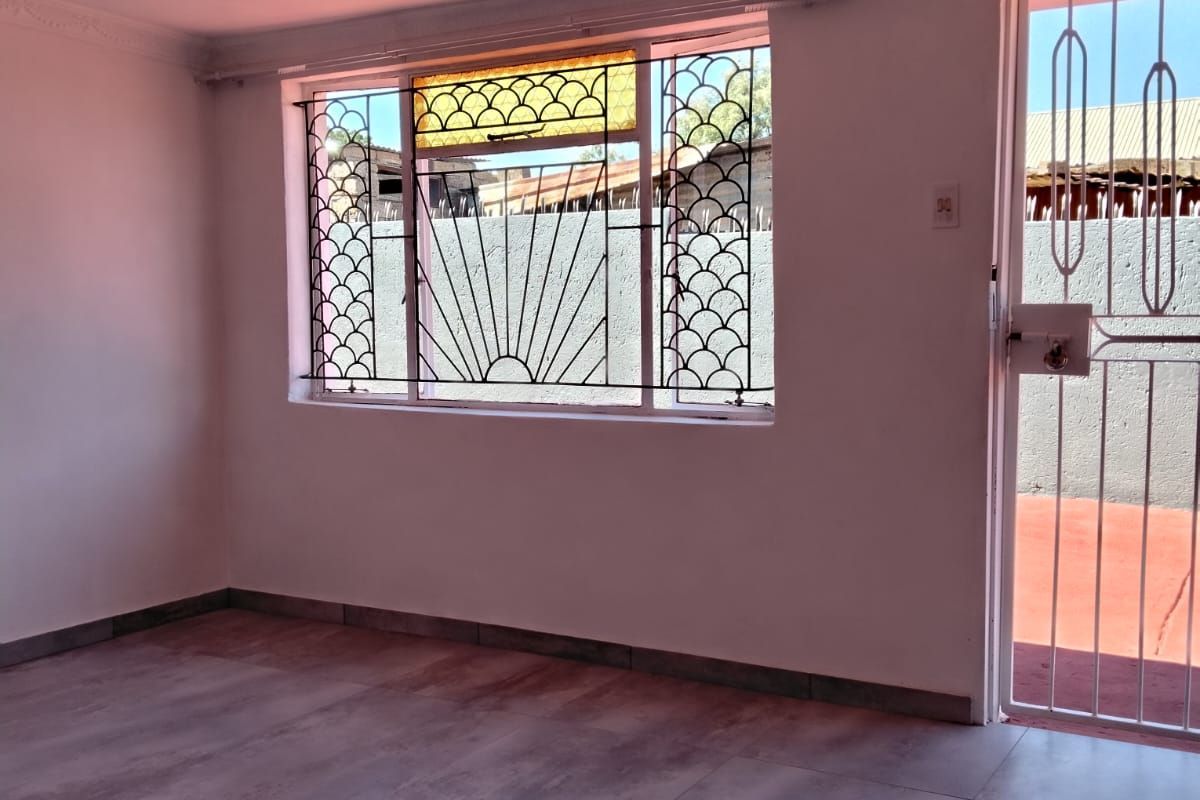 A bright, empty living room featuring large windows with decorative security bars, tiled flooring, and a secure metal entrance door.