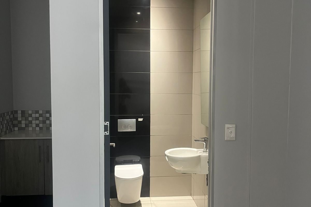 A modern, compact office restroom featuring a wall-mounted toilet, sleek black and beige tiling, and a wall-hung basin.